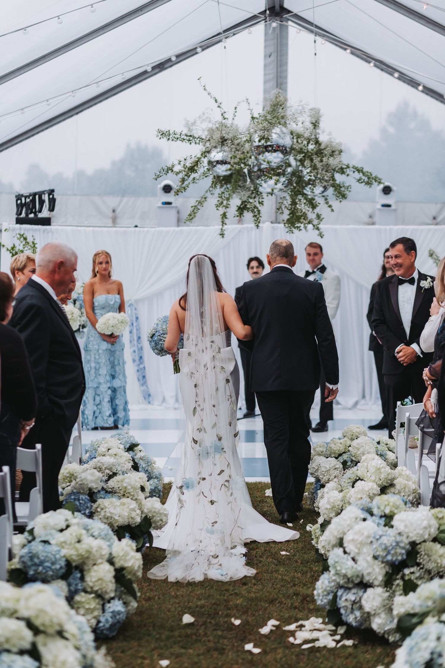 Bride walks down aisle lined with blue and white hydrangeas in a clear tent wedding ceremony with floral chandelier.