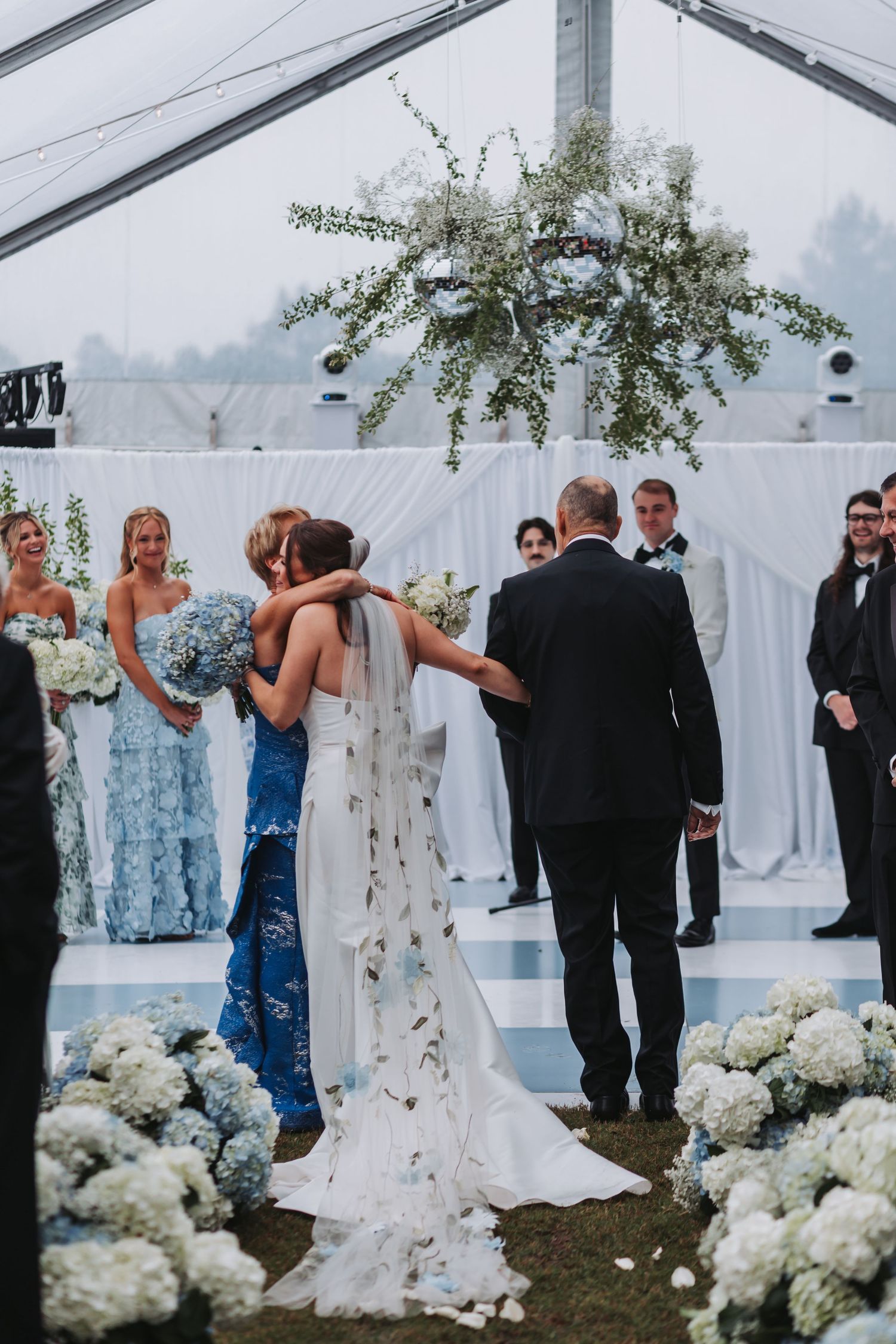 Bride embraces family member at altar during emotional wedding ceremony under floral chandelier in tent.