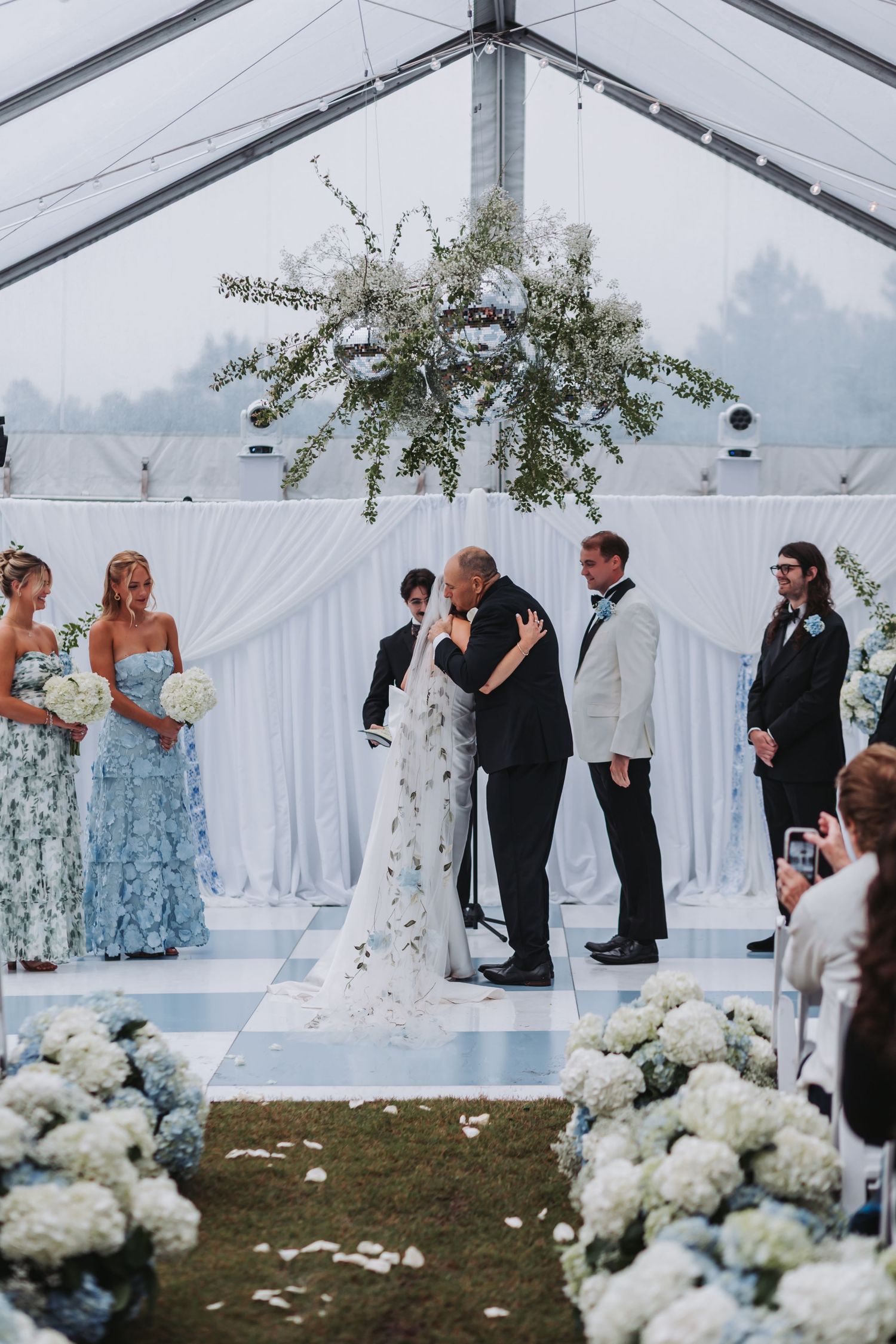 Bride and groom share first kiss at altar surrounded by wedding party under floral chandelier.