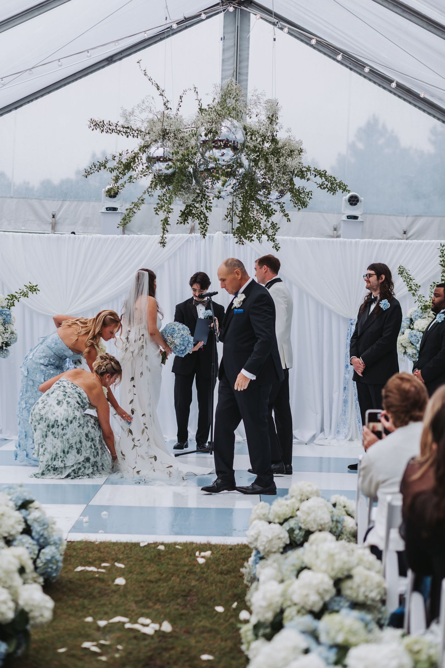 Wedding party gathers at altar as bride arrives in floral gown under hanging greenery installation in white draped tent.