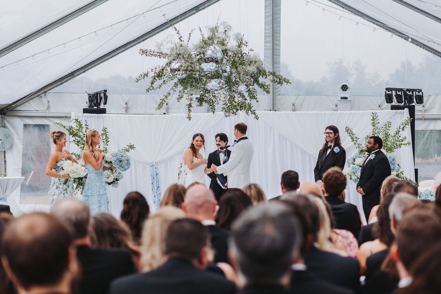 Wedding ceremony under clear tent with guests watching couple exchange vows at floral altar.