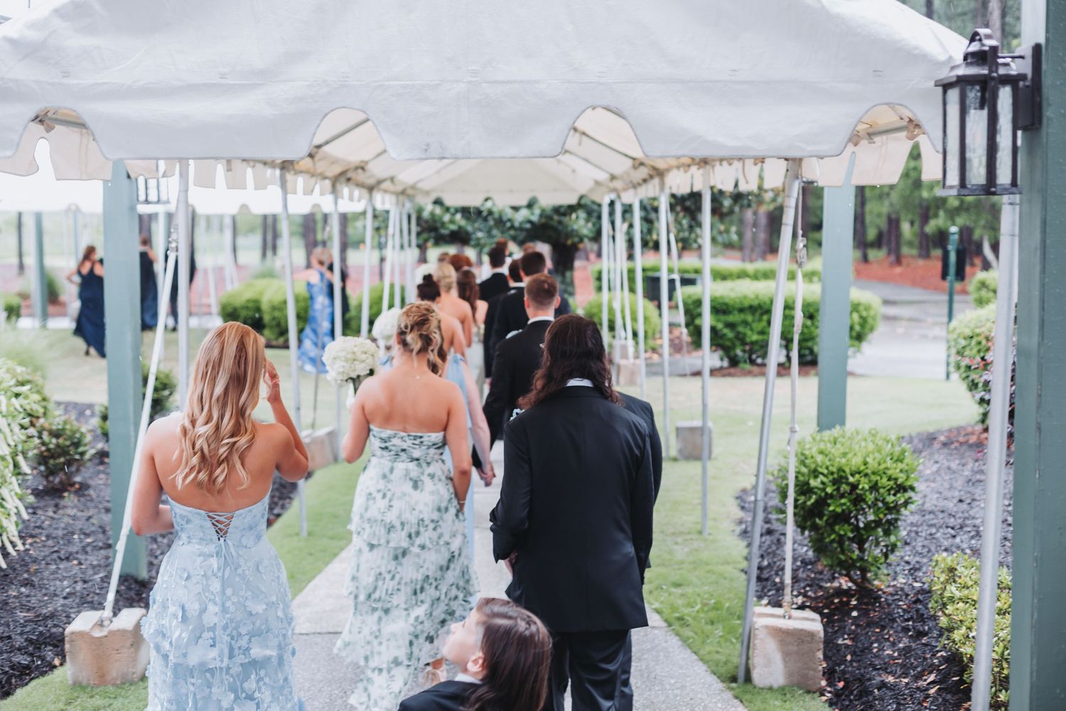 Wedding party walking down outdoor aisle under white tent canopy at garden venue ceremony.