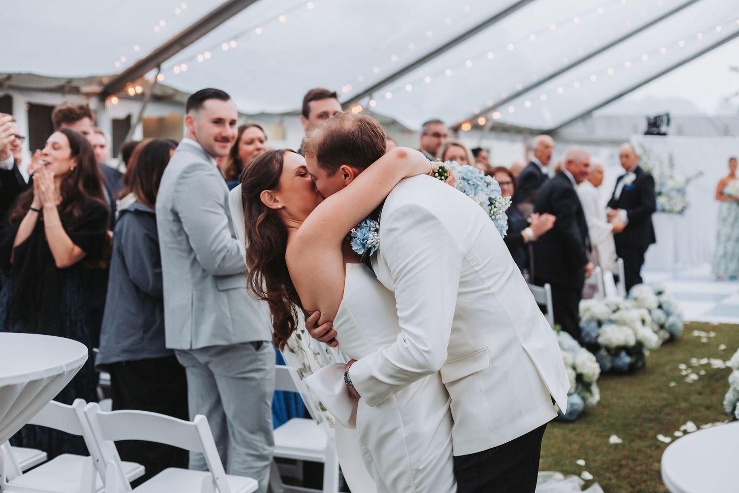 Bride and groom kiss as wedding guests cheer under a tent with string lights at outdoor reception.