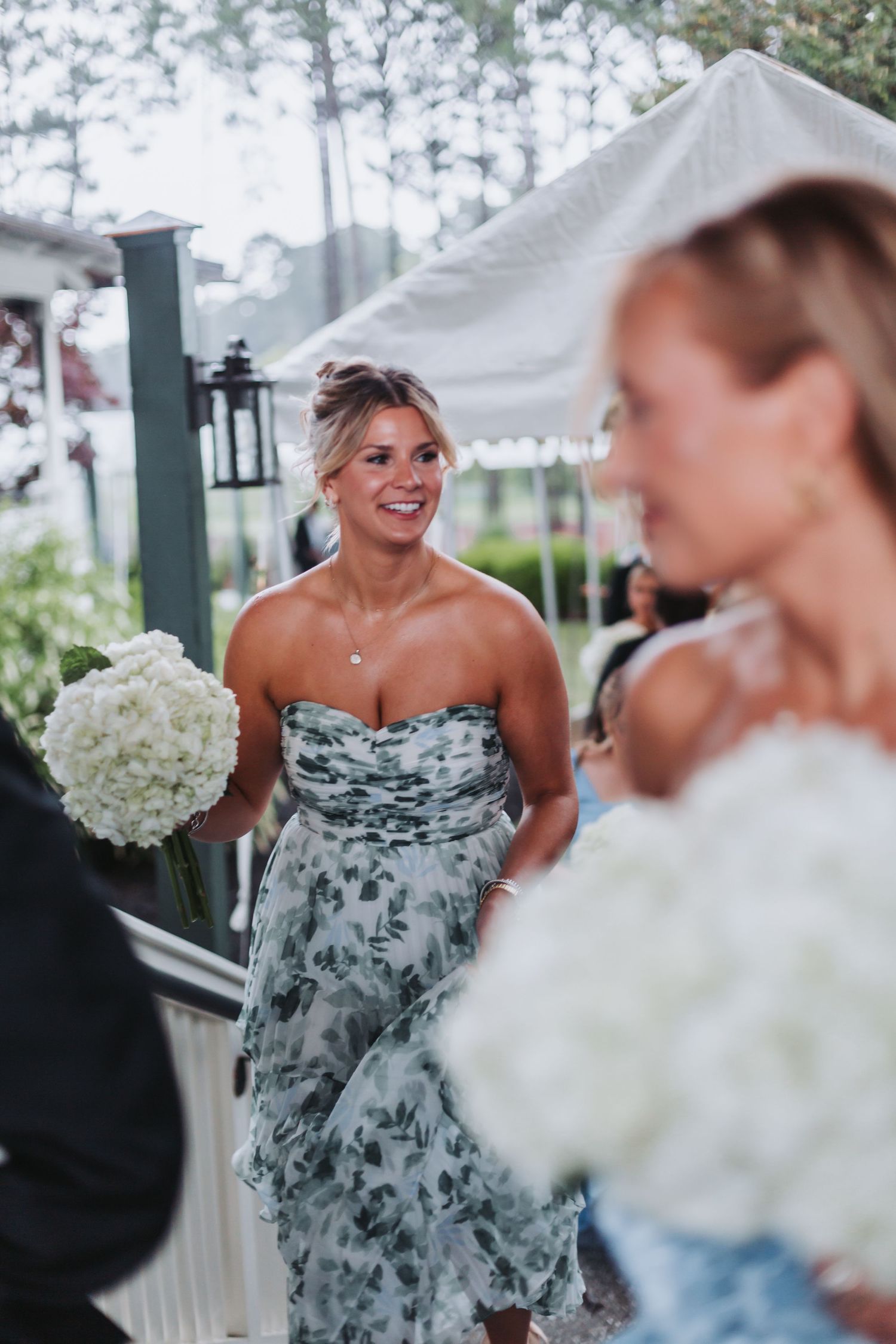 Smiling bridesmaid in strapless floral dress holds white hydrangea bouquet at outdoor wedding.