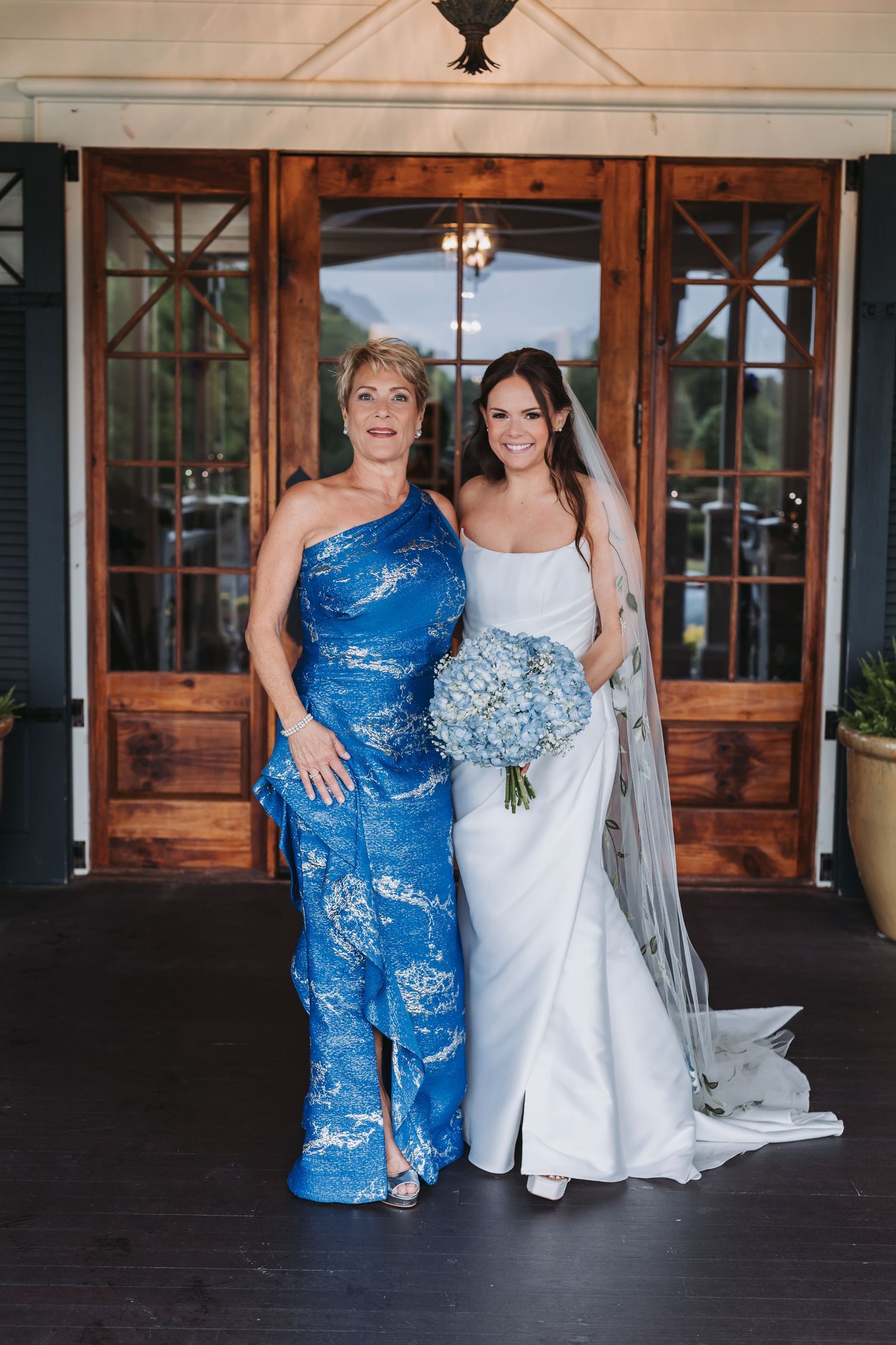 Bride in white gown holds blue hydrangea bouquet beside woman in blue floral jumpsuit before wooden doors.