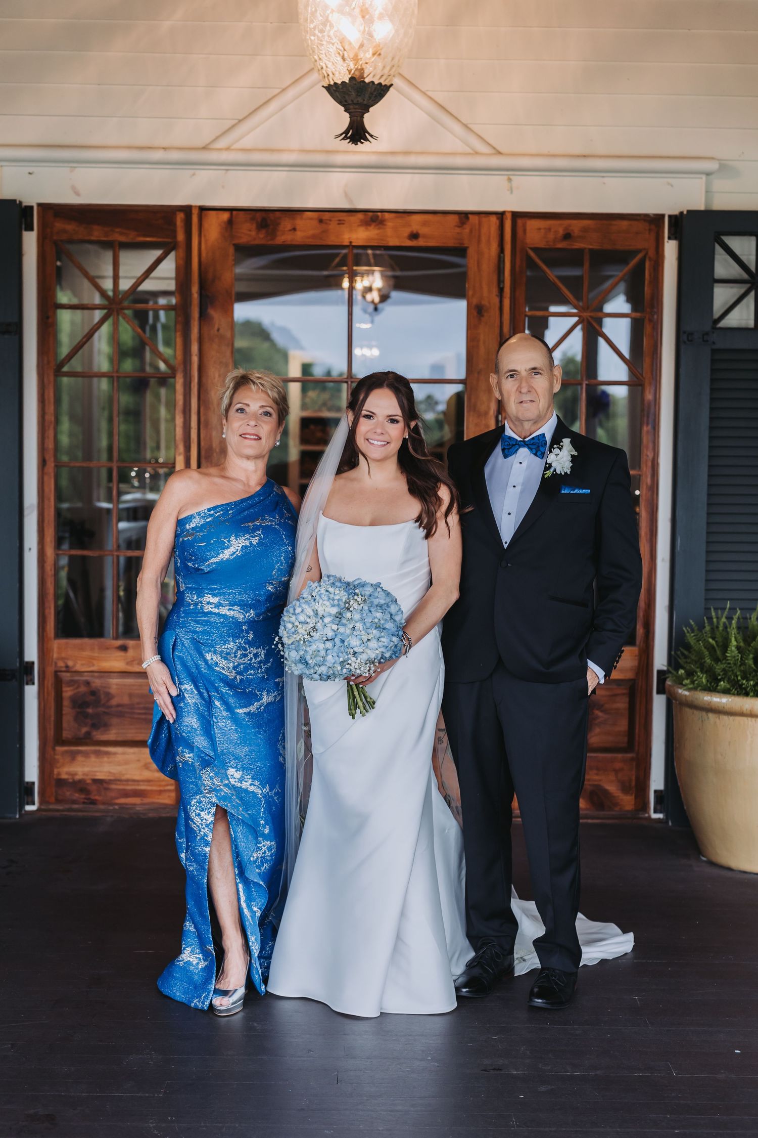 Bride with blue hydrangea bouquet stands between parents, mother in blue floral gown and father in tuxedo.