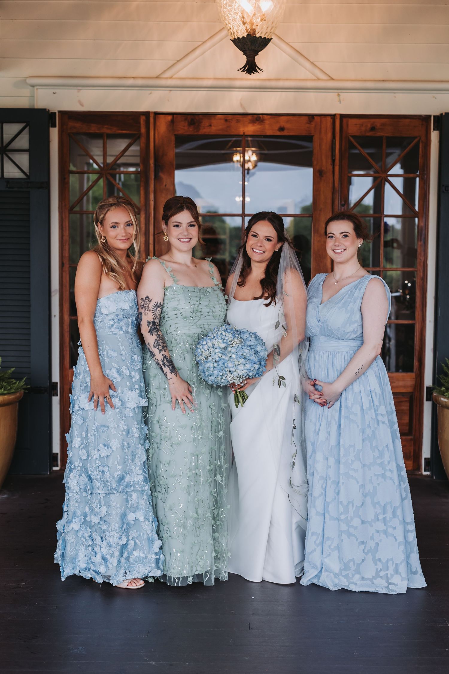 Bride in white gown poses with three bridesmaids in mismatched light blue dresses before wooden doors.