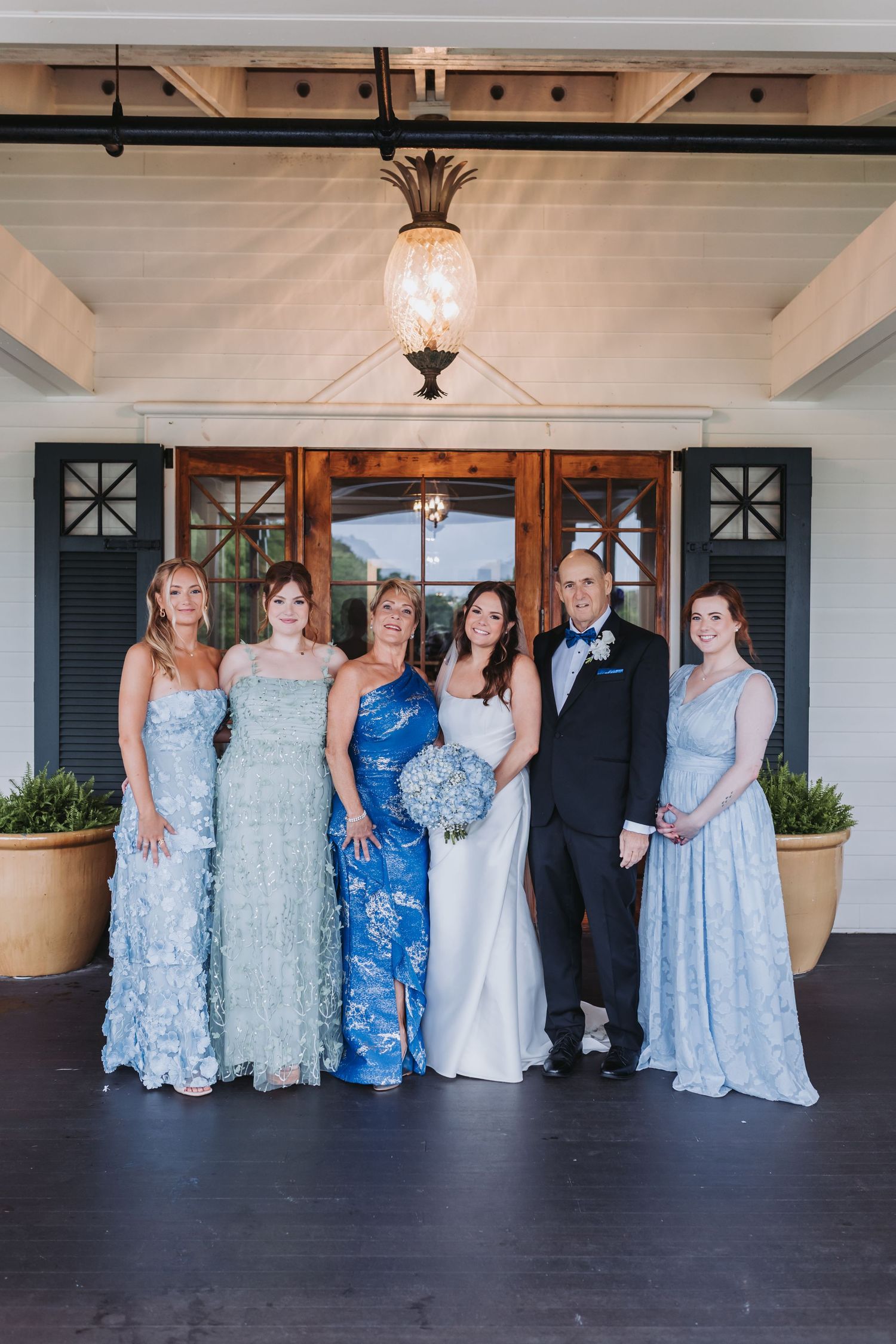 Bride and groom pose with family in blue formal gowns before wooden doors at wedding venue.