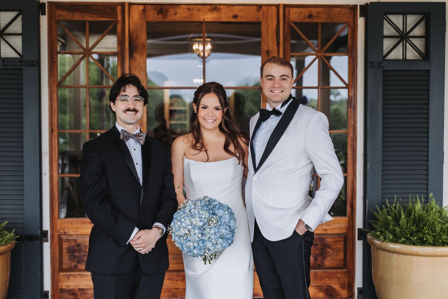 Bride in white strapless gown holds blue bouquet flanked by two groomsmen in black and white tuxedos.