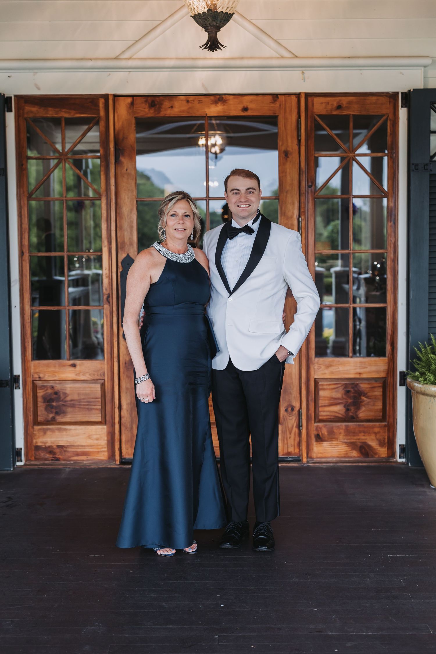 Groom in white tuxedo jacket posing with mother in navy gown at venue entrance.