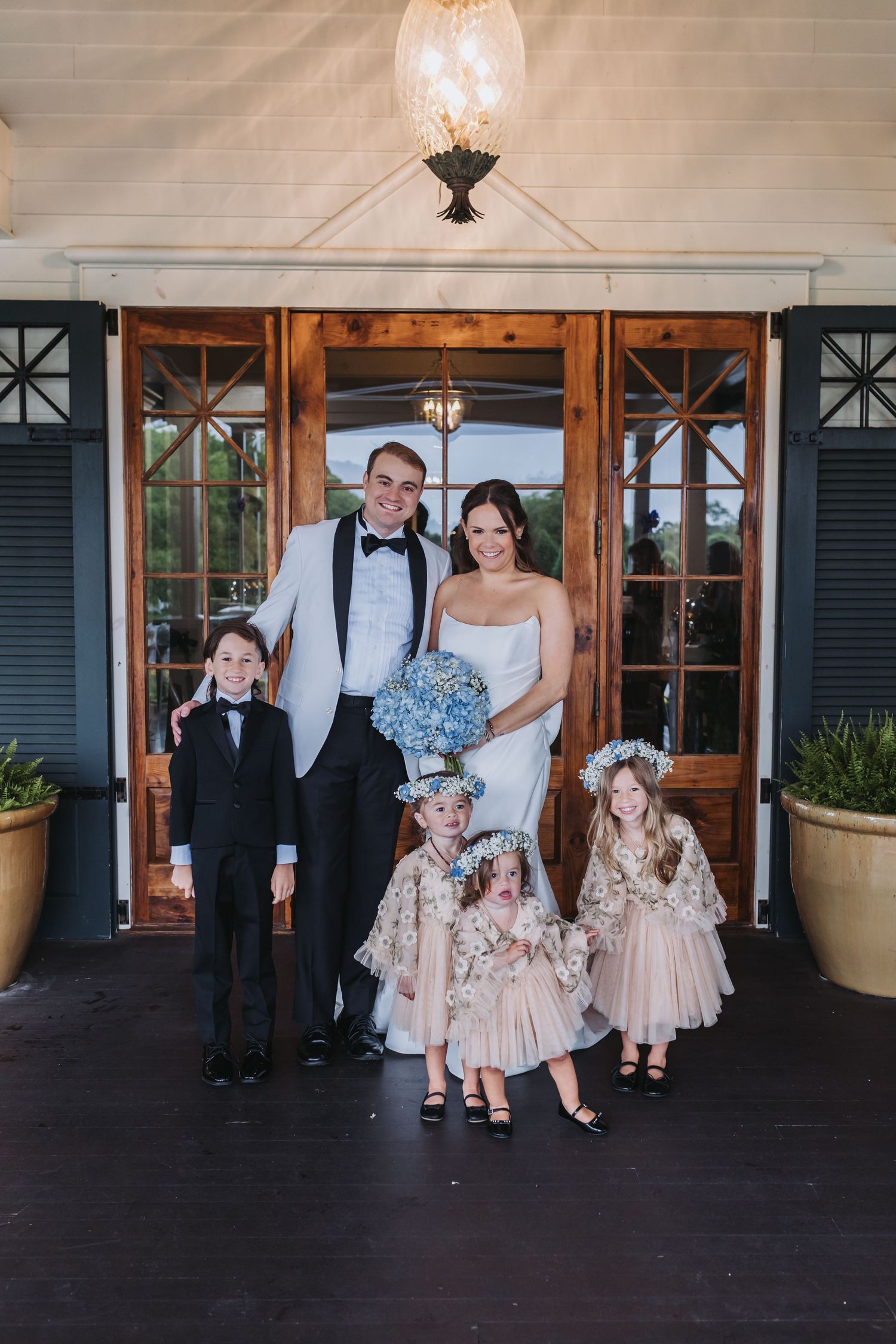 Bride and groom pose with flower girls and ring bearer in front of wooden doors at elegant wedding venue.