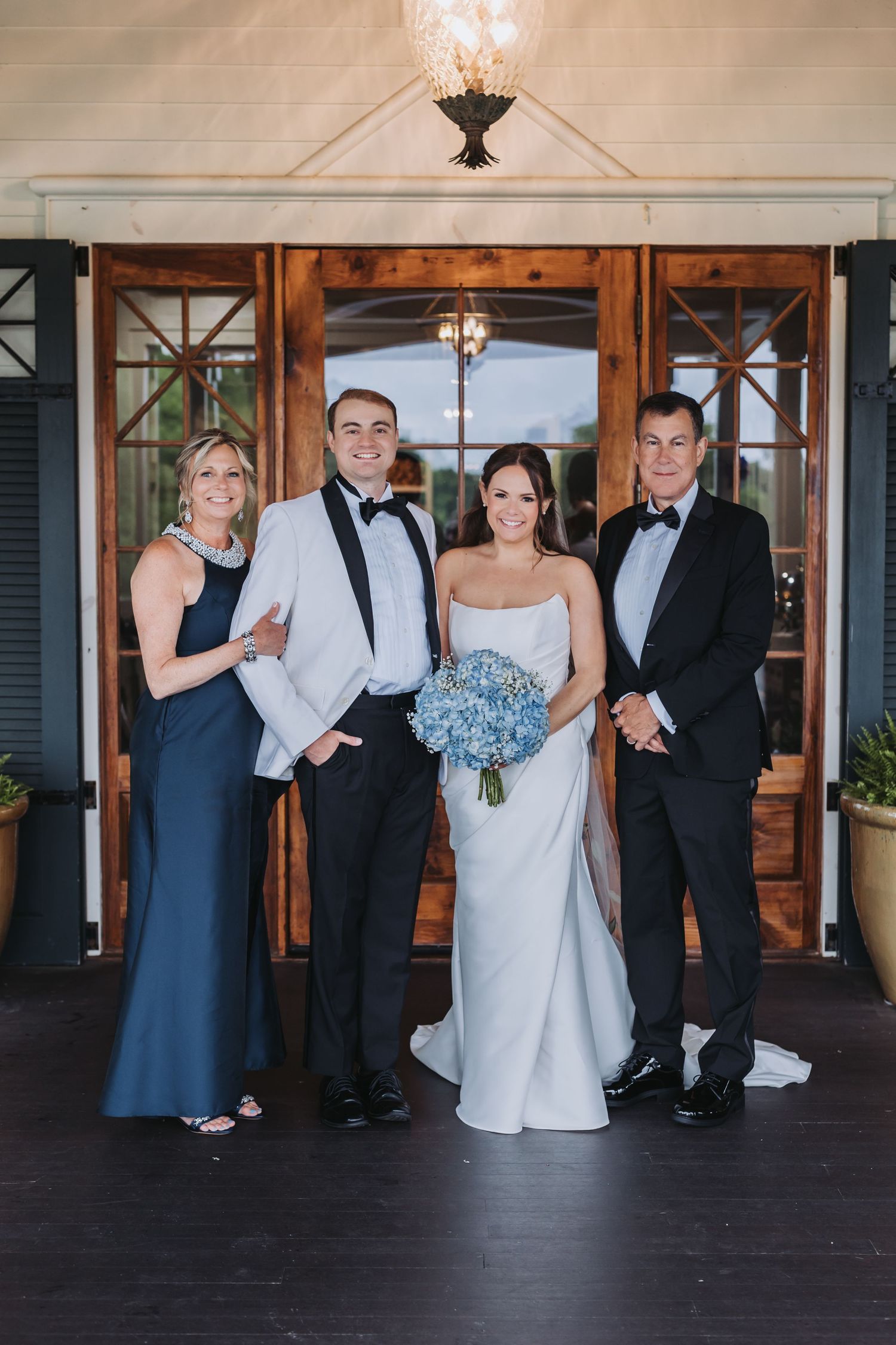 Bride holding blue hydrangea bouquet poses with groom and parents in formal attire at wedding.