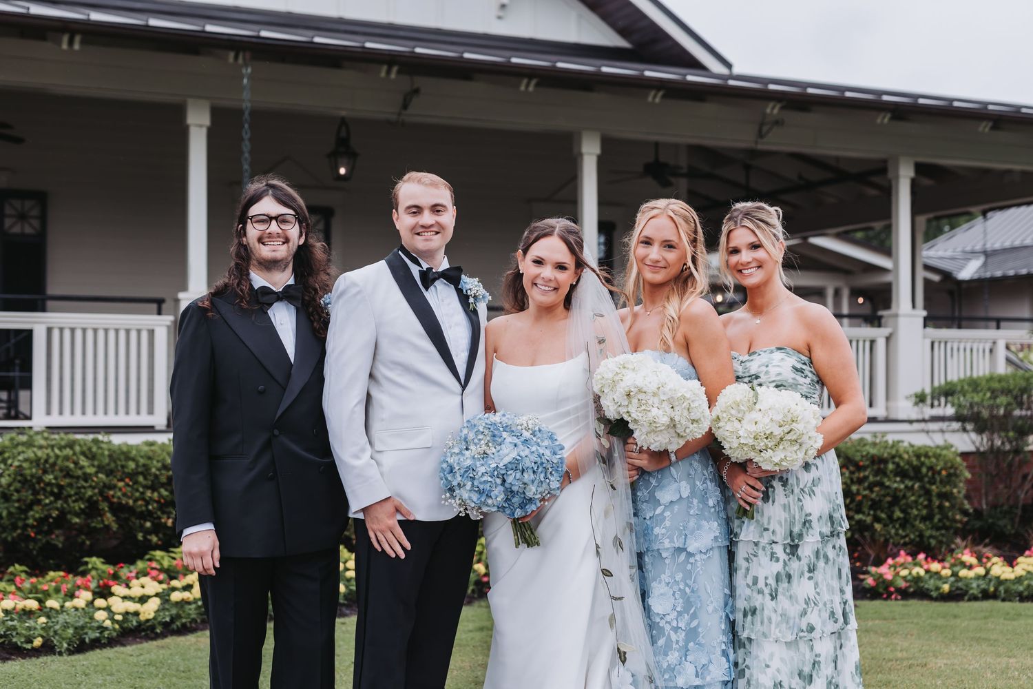 Wedding party of five posing outdoors, bride in white gown with blue hydrangea bouquet at elegant venue.