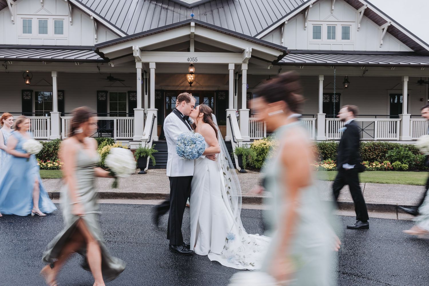 Bride and groom kiss outside elegant venue while wedding party walks past in blur.