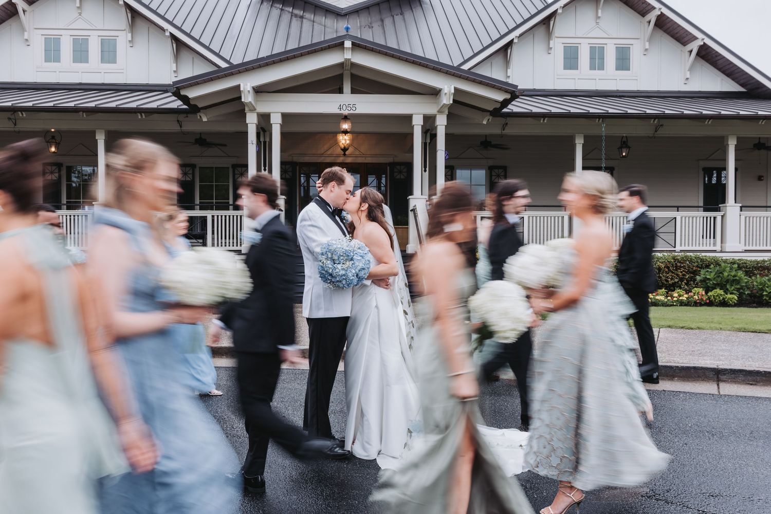 Bride and groom share a kiss as wedding party in blue gowns moves past them.