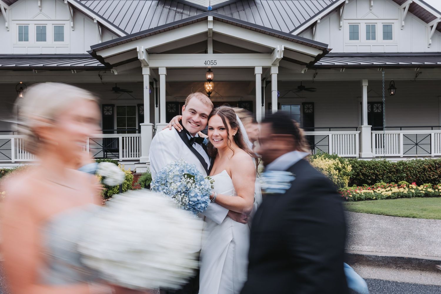 Bride and groom embrace outside elegant venue while wedding party passes in foreground.