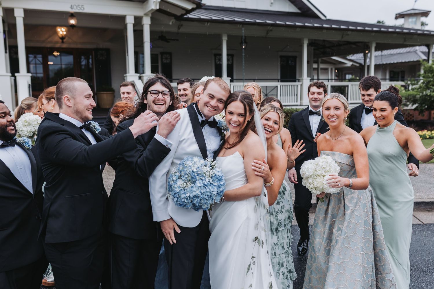 Bride and groom laugh with cheerful wedding party gathered closely outside elegant venue.