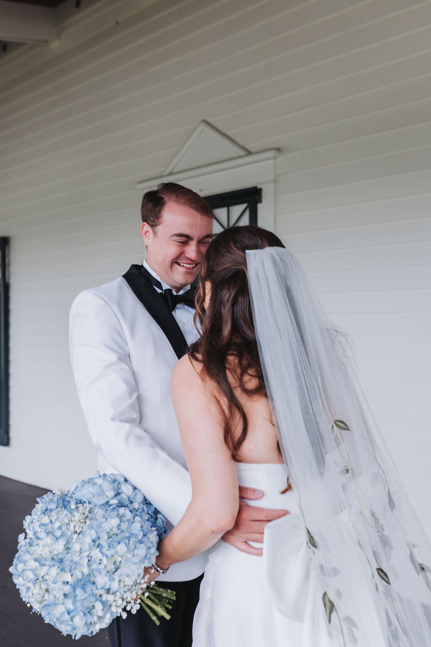 Smiling groom in white tuxedo embracing bride holding blue hydrangea bouquet at wedding.