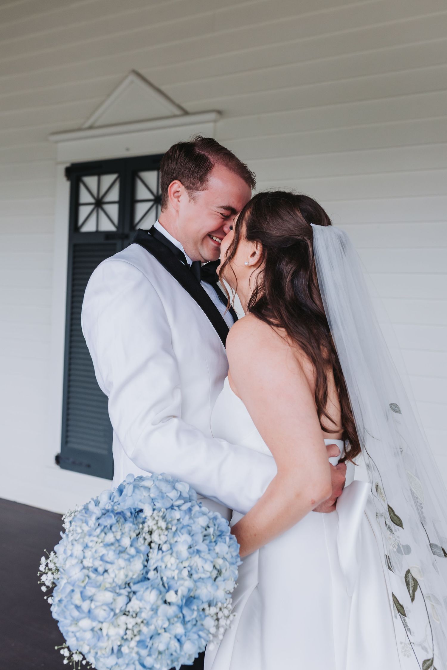 Joyful bride and groom share intimate moment, groom holding blue hydrangea bouquet outside white chapel.