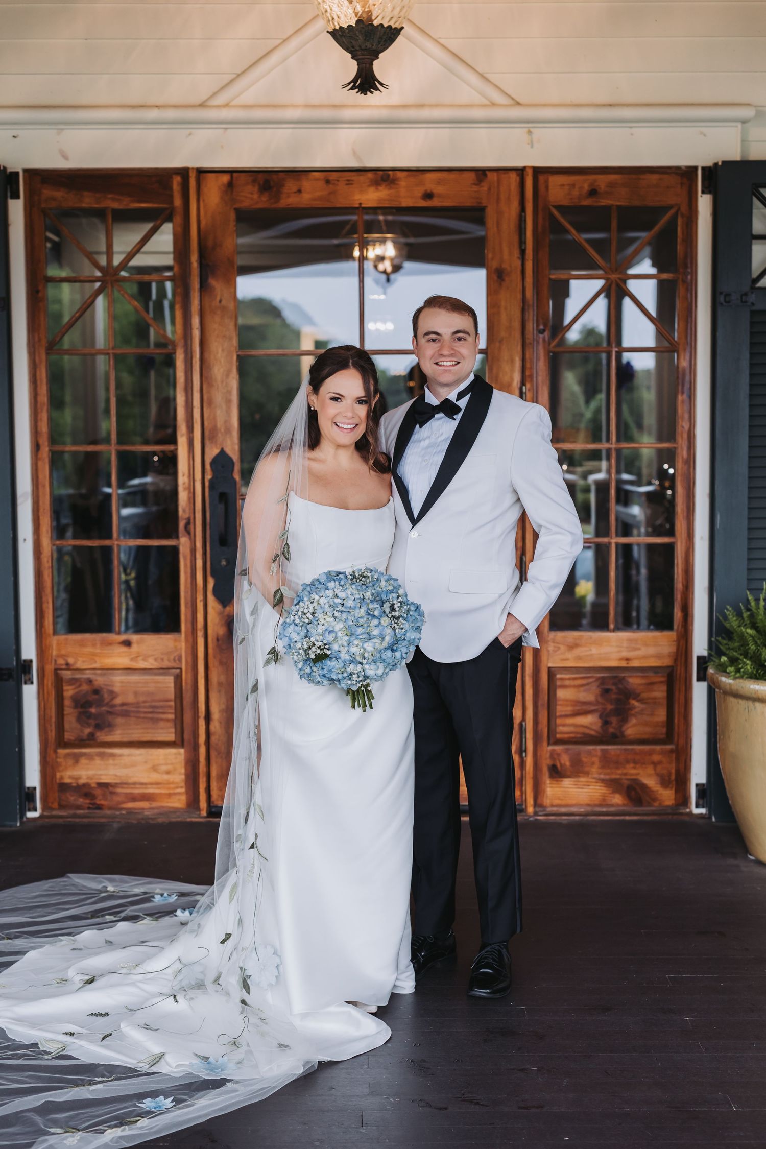 Bride in white gown with floral veil and groom in white jacket pose at wooden venue doors.
