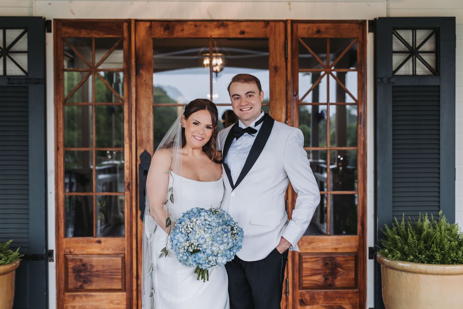 Bride holding blue hydrangea bouquet stands with groom in white tuxedo at wooden venue doors.
