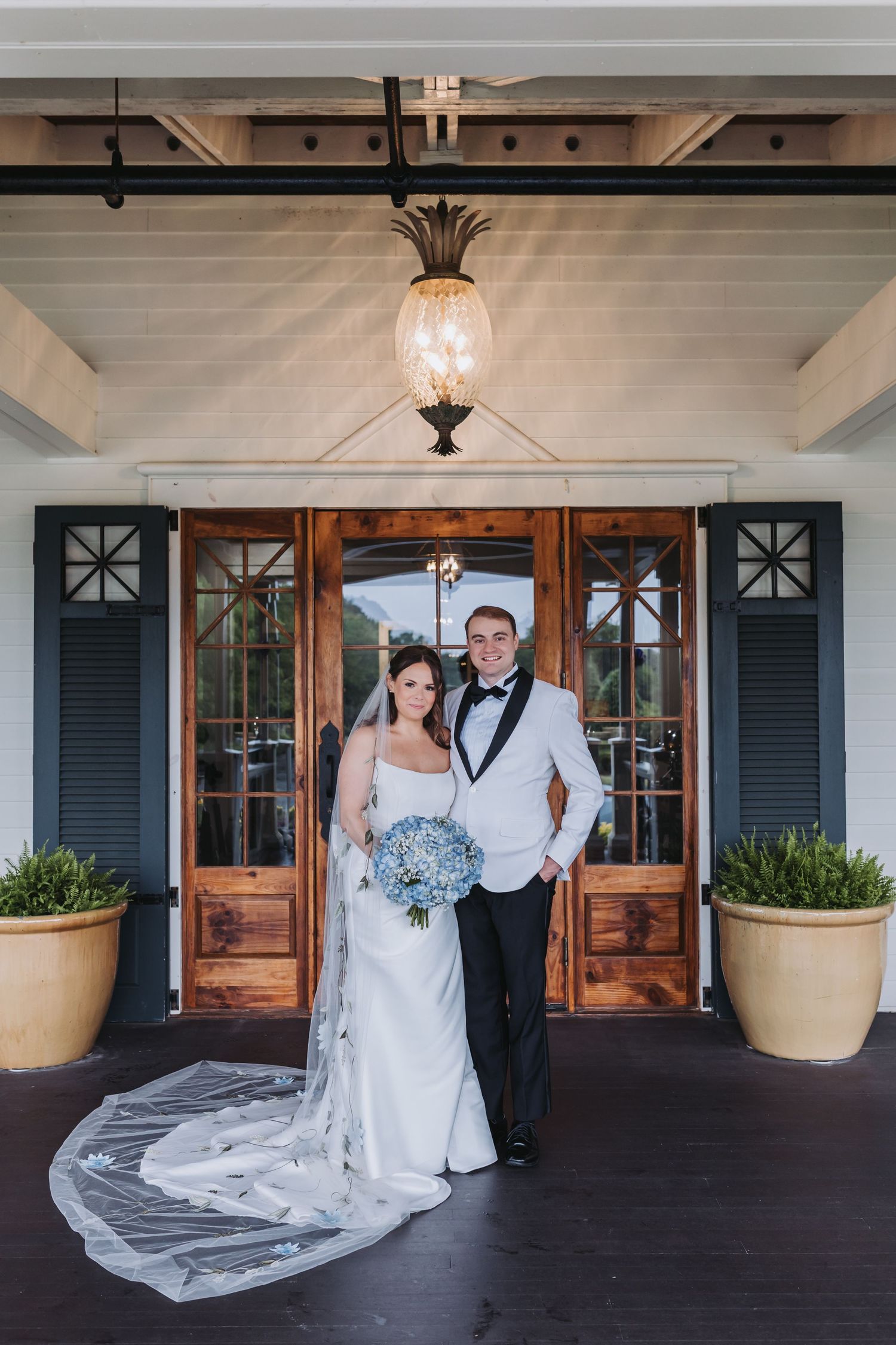Bride and groom stand under pineapple chandelier holding blue hydrangea bouquet at venue entrance.