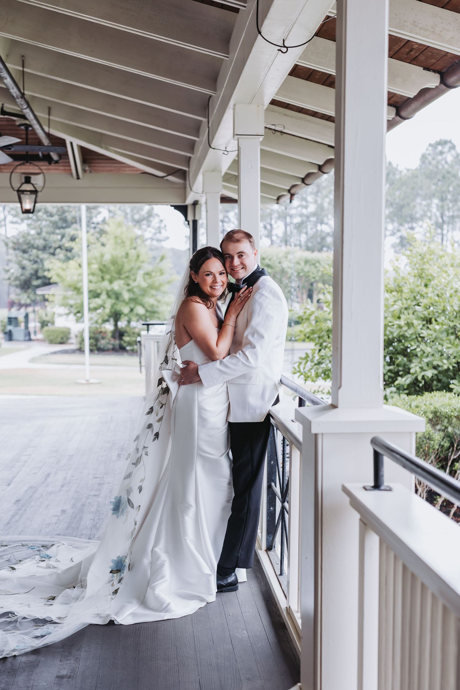 Bride and groom embrace on a white colonial porch, bride in strapless gown with floral train, groom in white tuxedo jacket.