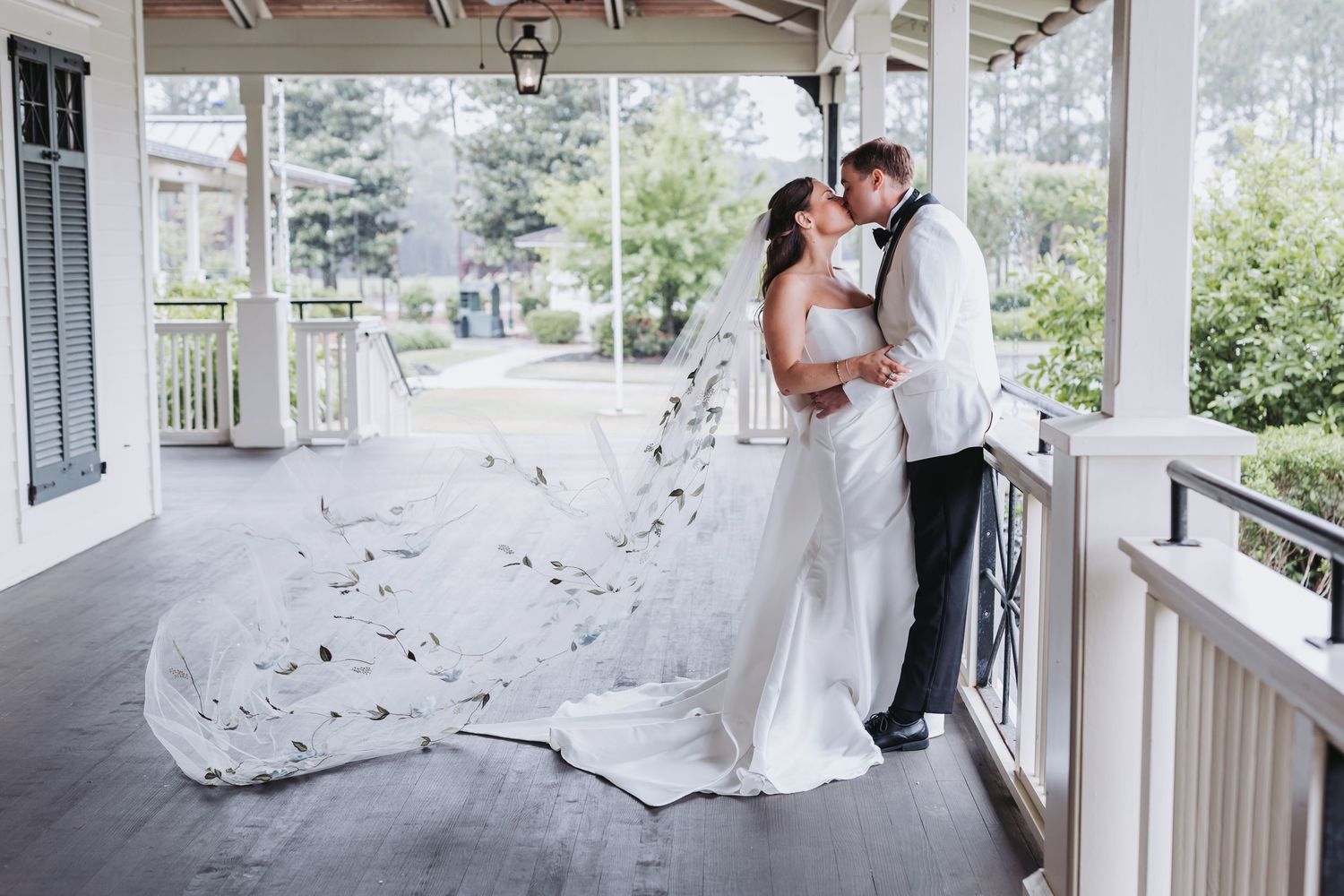 Bride and groom kiss on porch as cathedral veil with floral details flows dramatically in wind.