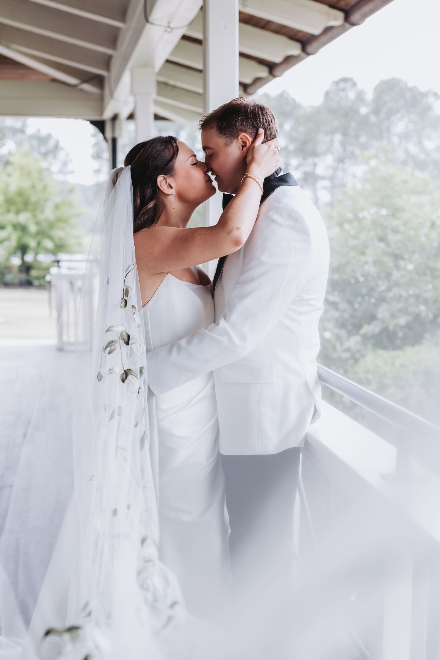 Bride and groom sharing a romantic kiss on a white porch with veil flowing.