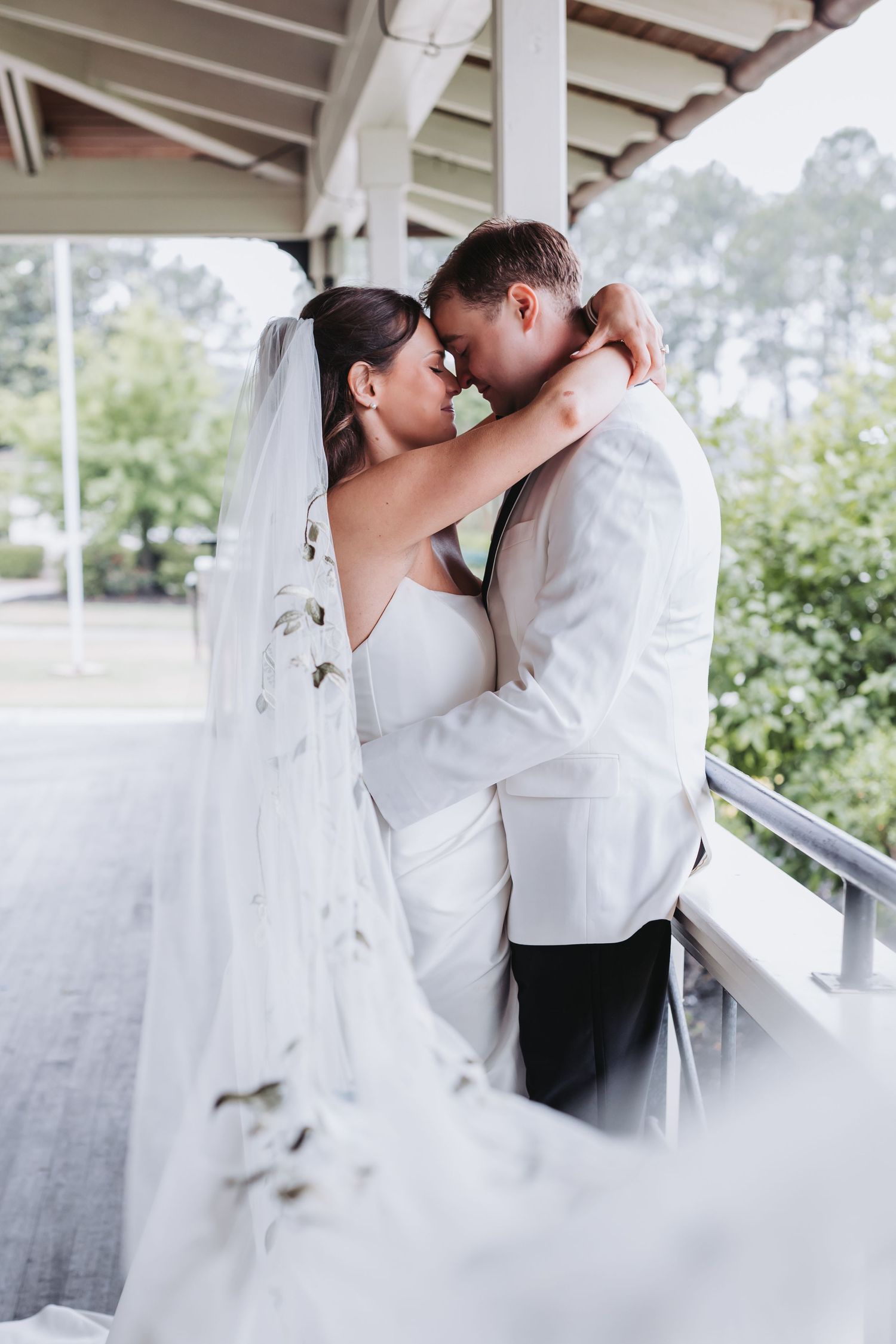 Bride and groom sharing romantic almost-kiss moment on porch, bride wearing long veil with floral details.
