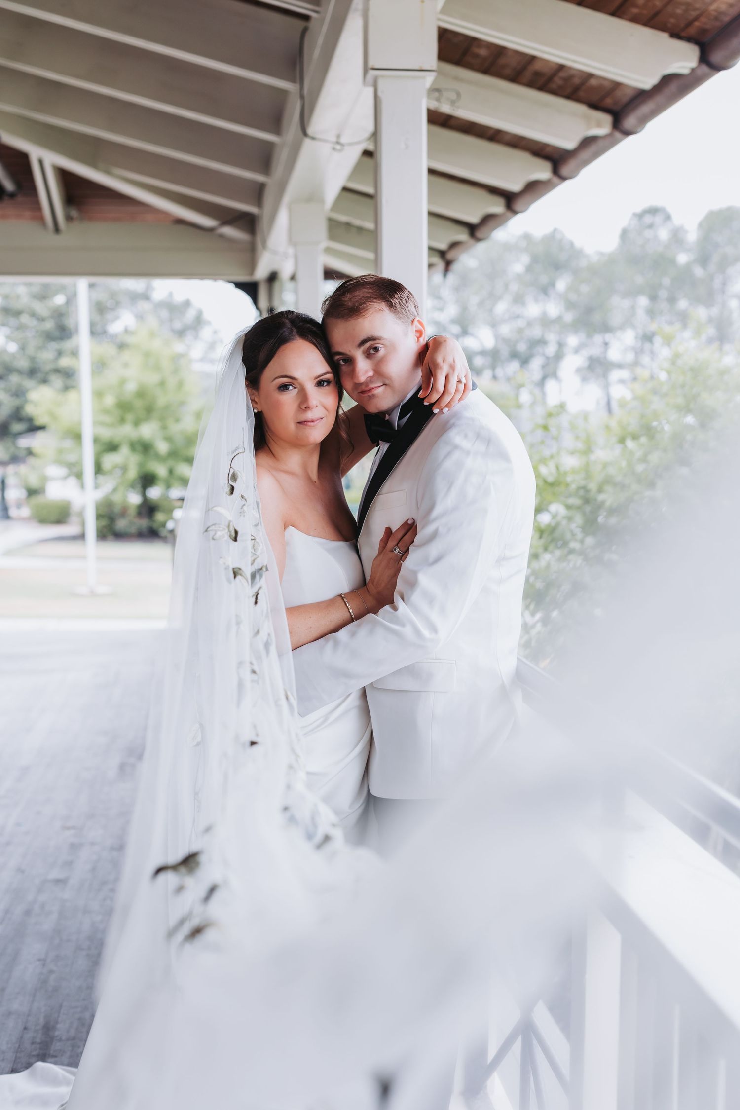 Bride and groom in white wedding attire embrace on a decorated porch, with lush greenery in the background.