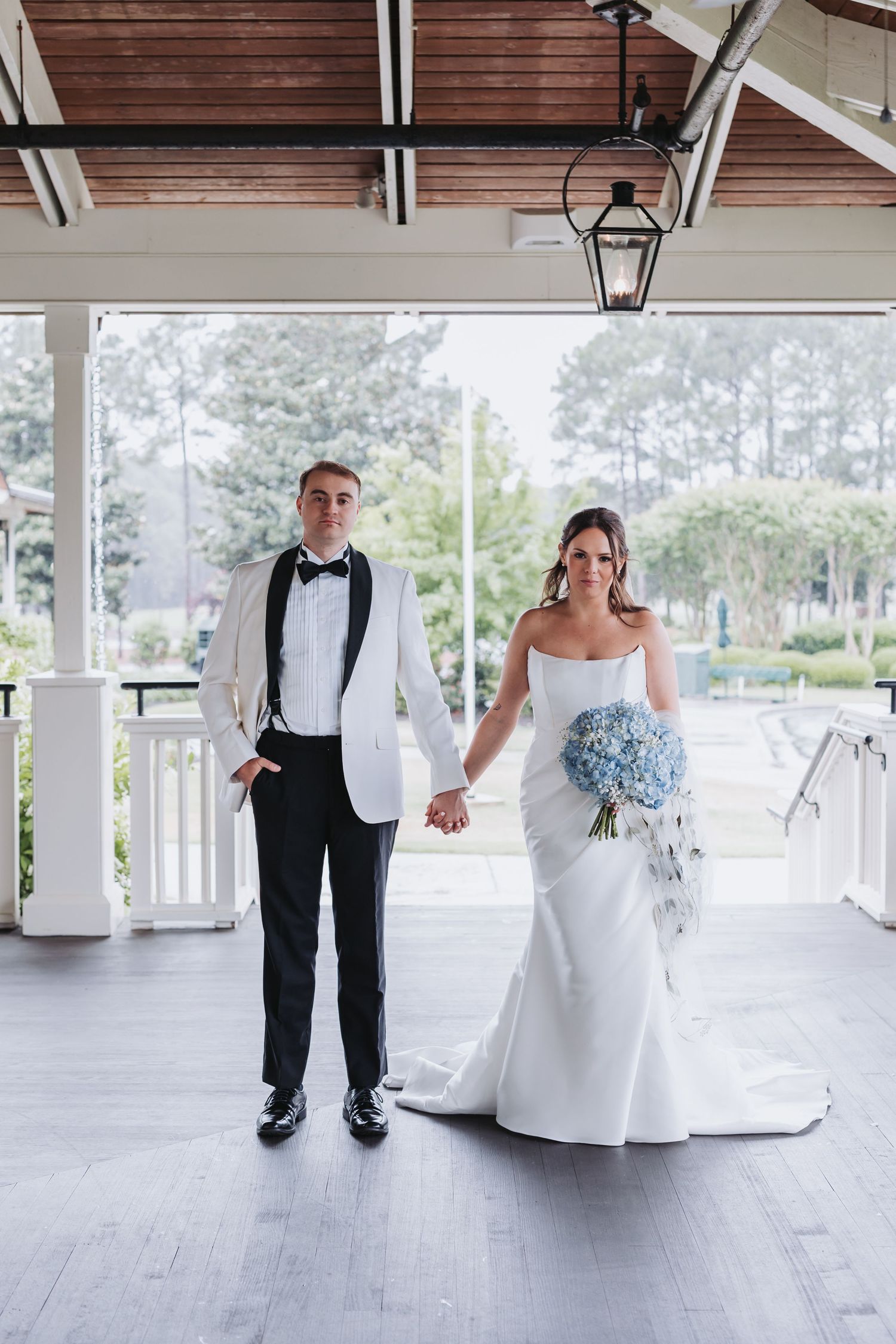 Elegant bride and groom hold hands on covered porch, bride carrying stunning blue hydrangea bouquet.