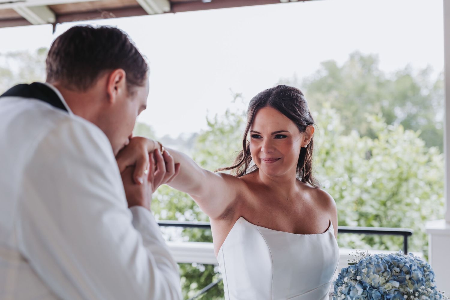 Radiant bride smiles as groom kisses her hand on outdoor porch, holding blue hydrangea bouquet.