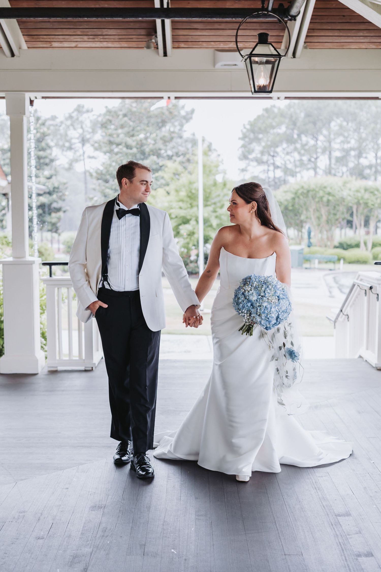 Wedding couple walks hand-in-hand on porch, bride holding blue hydrangea bouquet in strapless white gown, groom in white tuxedo.