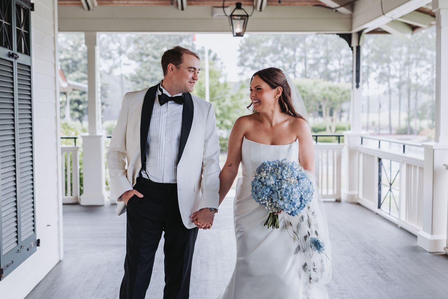 Bride and groom in white tuxedo hold hands walking on porch, bride carrying blue hydrangea bouquet.