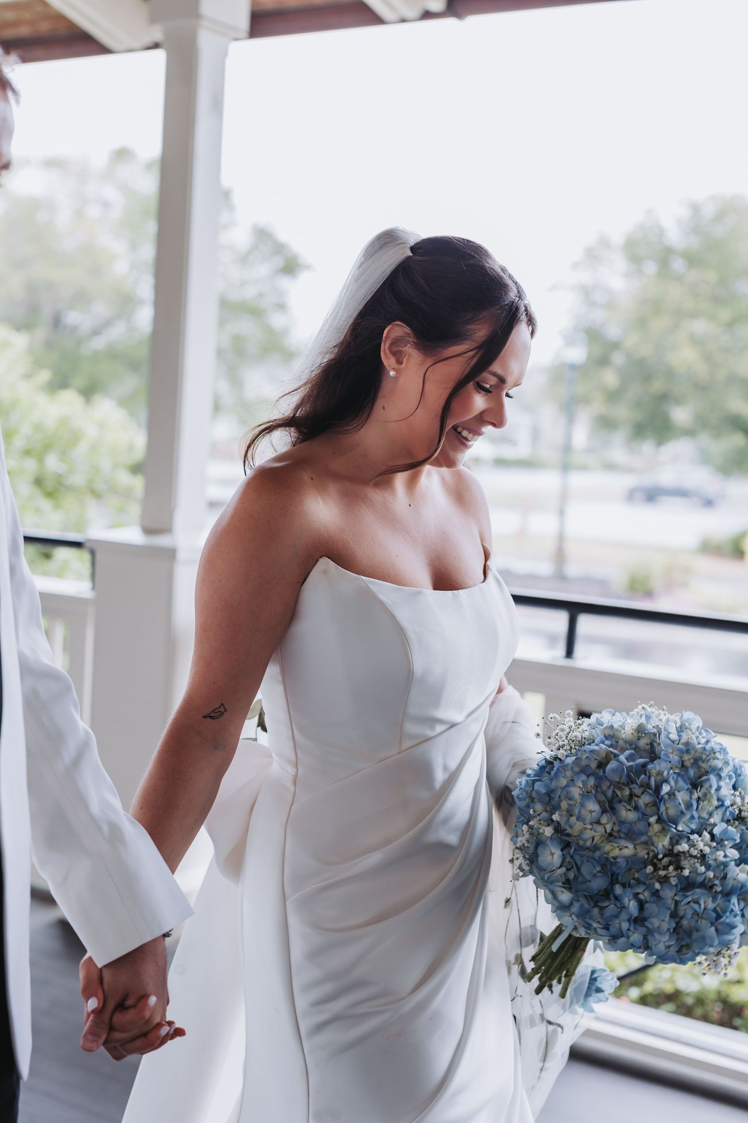 Smiling bride in strapless white gown holds blue hydrangea bouquet while holding hands on a porch.