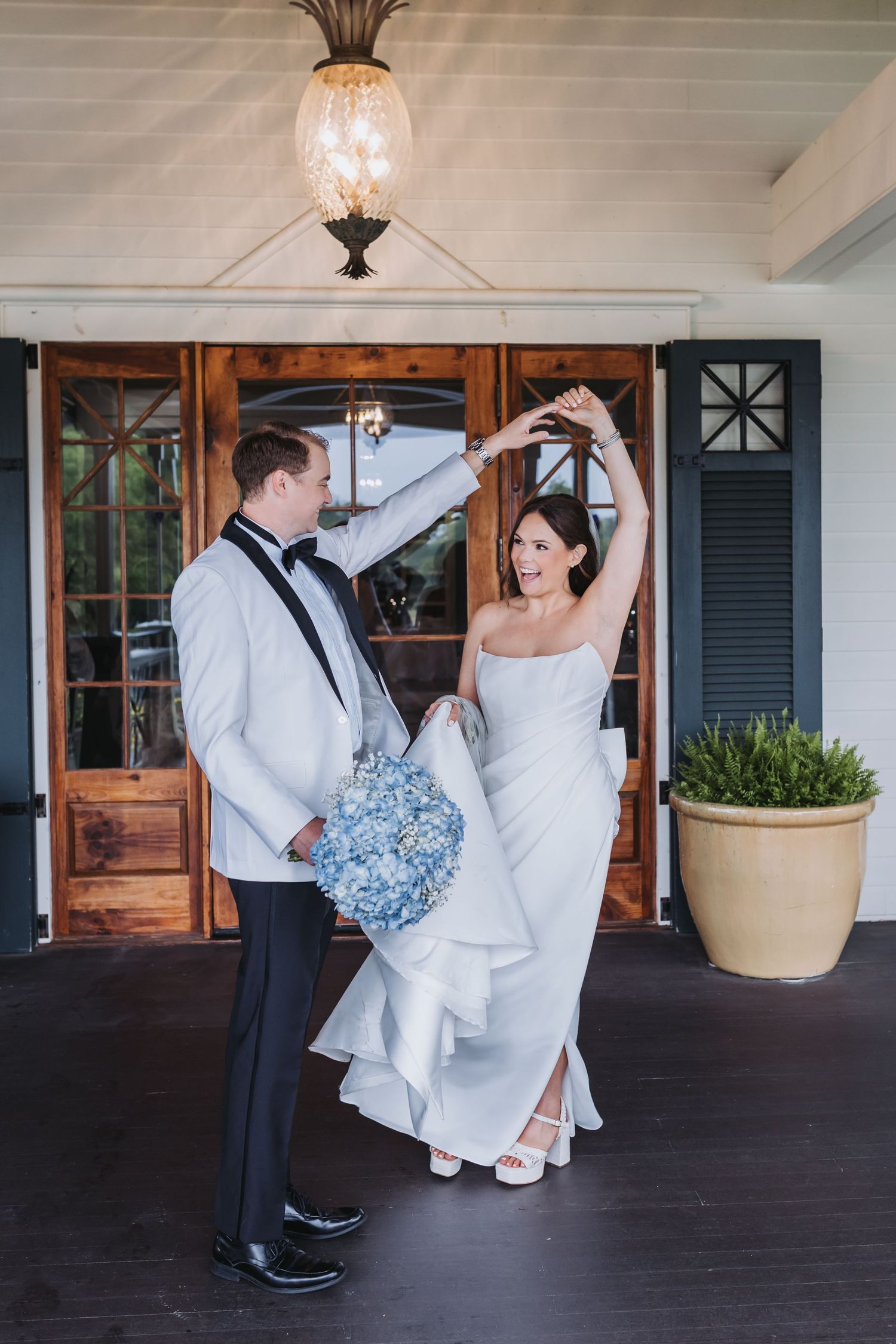 Groom in white tuxedo twirling bride in strapless gown on venue porch during wedding portraits.