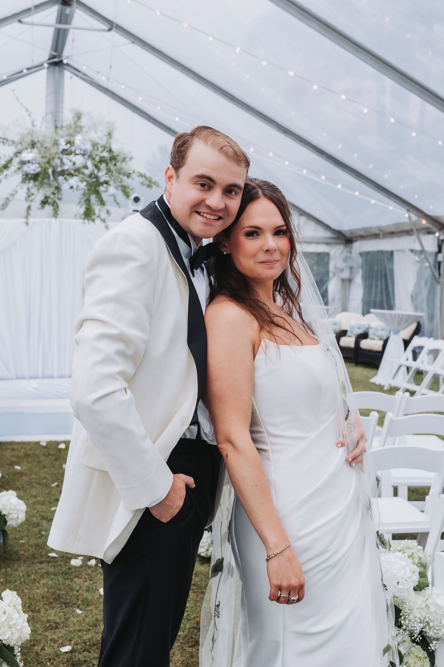 Bride and groom pose together under a white tent, groom in white tuxedo jacket with black bow tie.