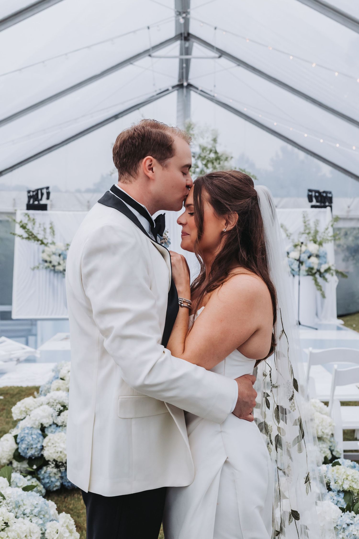 Bride and groom sharing tender kiss under clear wedding tent surrounded by blue and white hydrangeas.