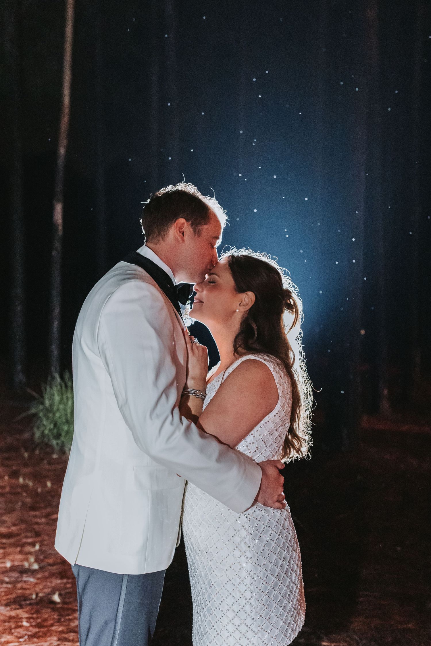 Bride and groom share a romantic kiss under a starry night sky surrounded by trees at their outdoor wedding.