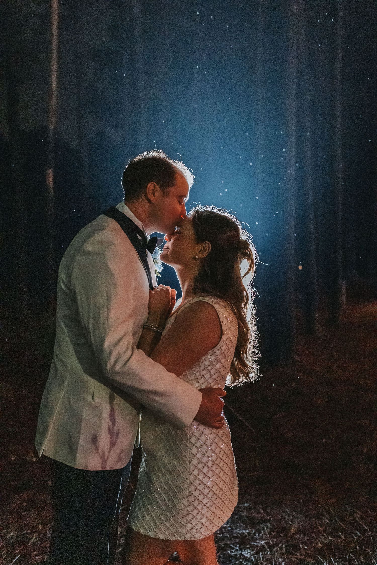 Bride and groom sharing romantic kiss outdoors at night in forest with starry backdrop.