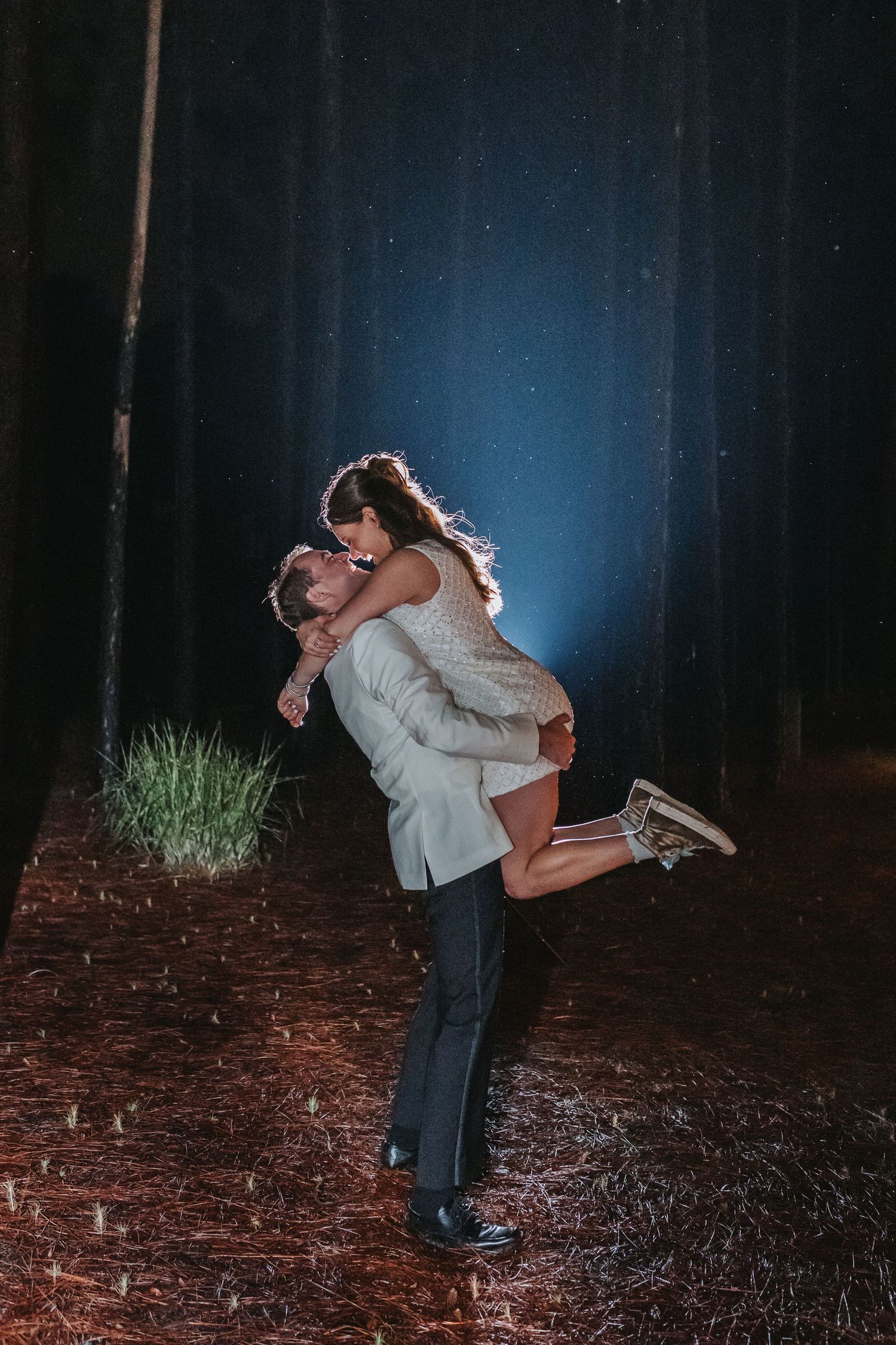 Groom lifts bride in sequin dress during romantic nighttime outdoor wedding photo in forest setting.