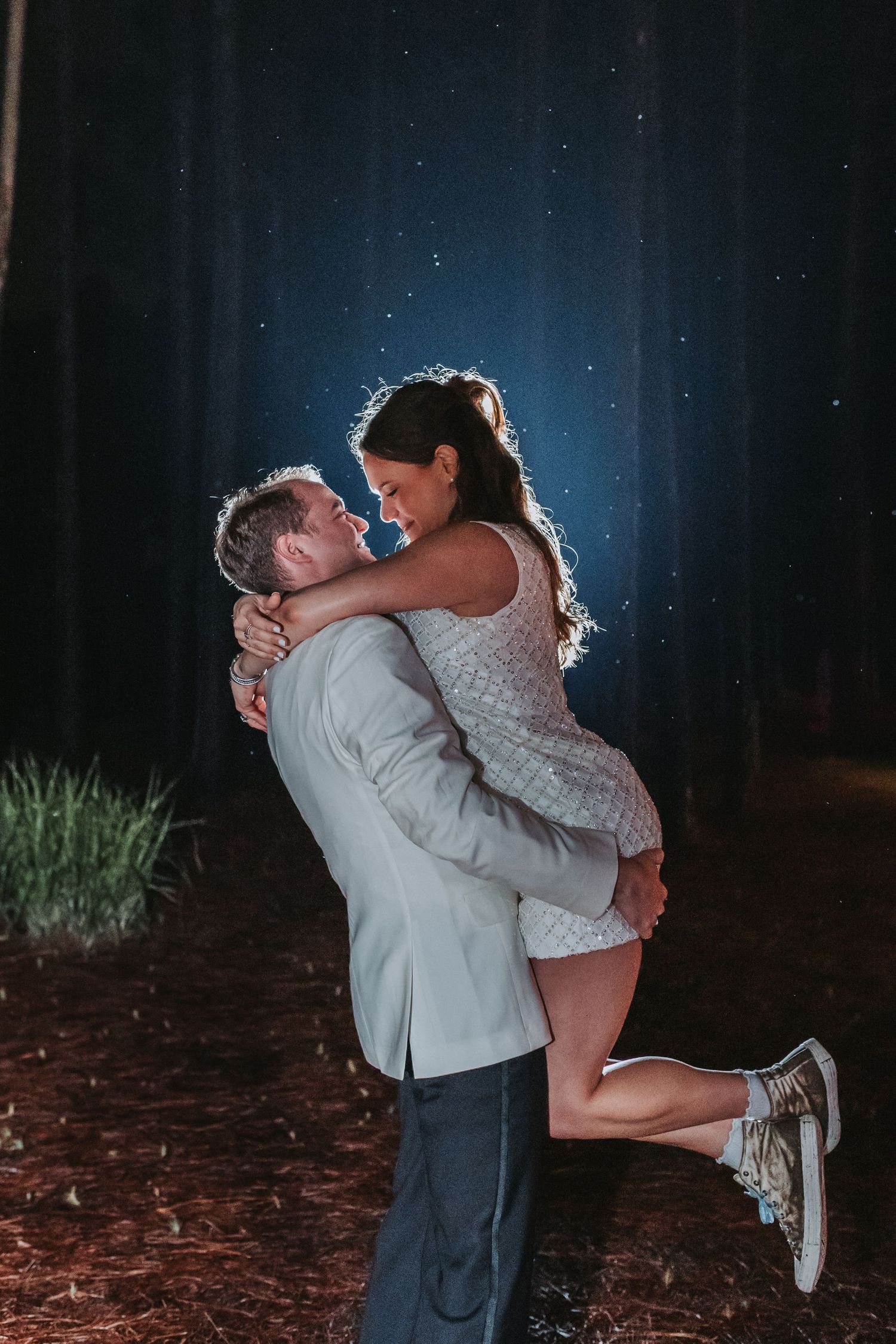 Groom lifting bride in white sequin dress joyfully in illuminated forest at night.