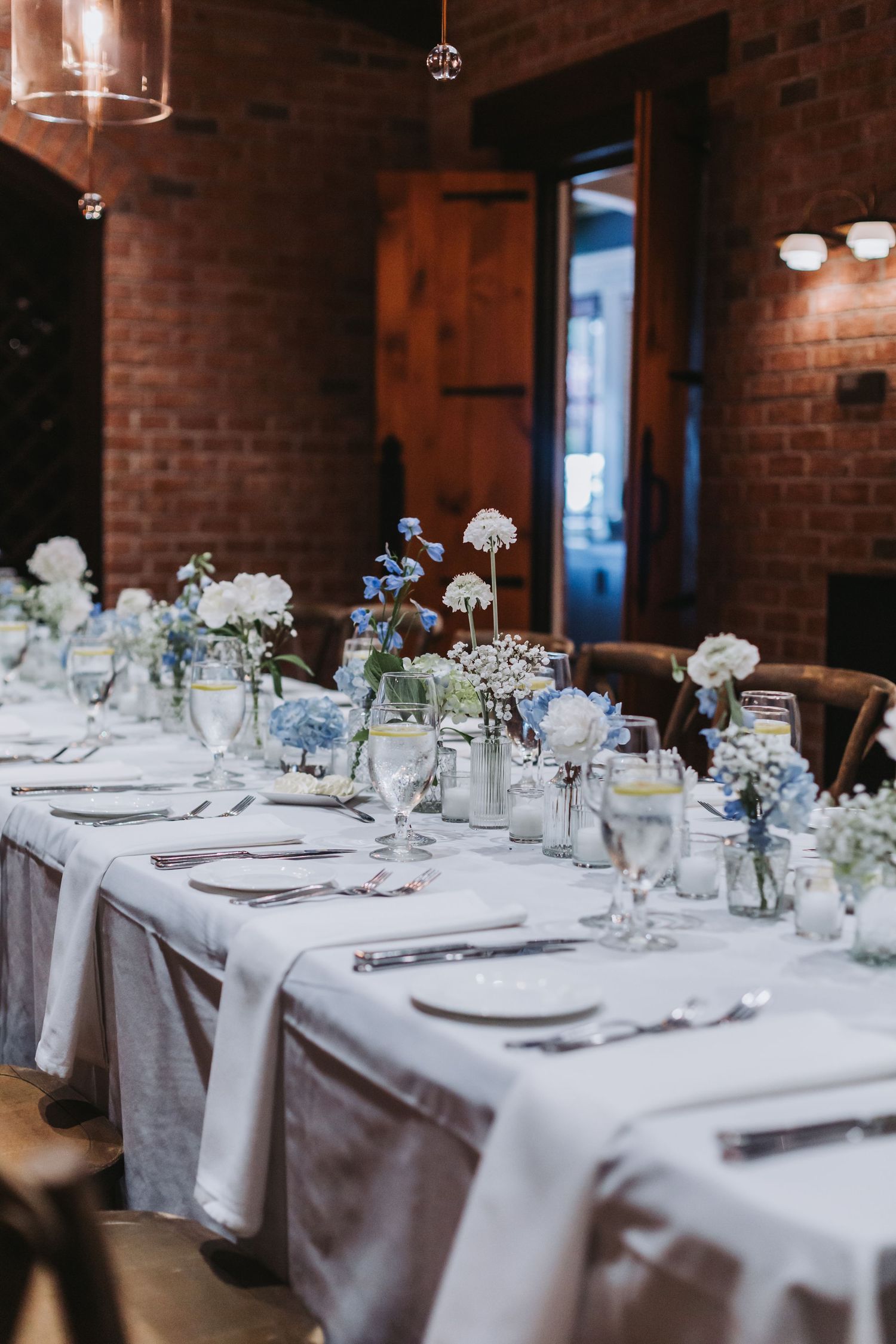 Elegant wedding reception table set with white linens, blue and white hydrangea centerpieces in a brick-walled venue.
