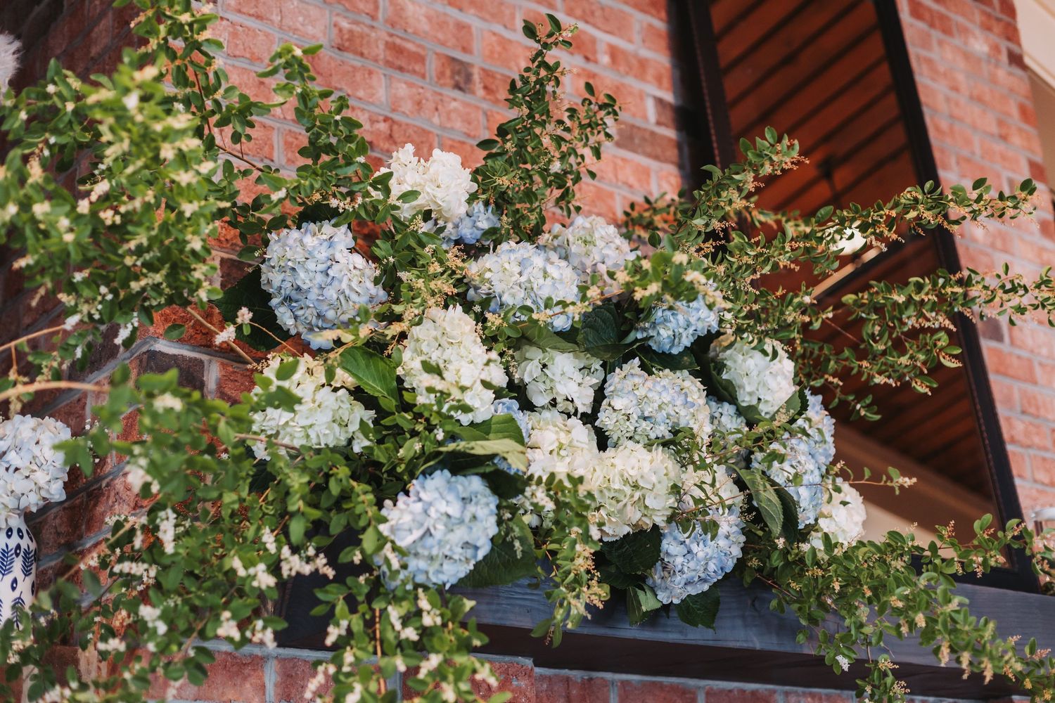 Blue and white hydrangea floral arrangement with greenery displayed on a brick wall window ledge.