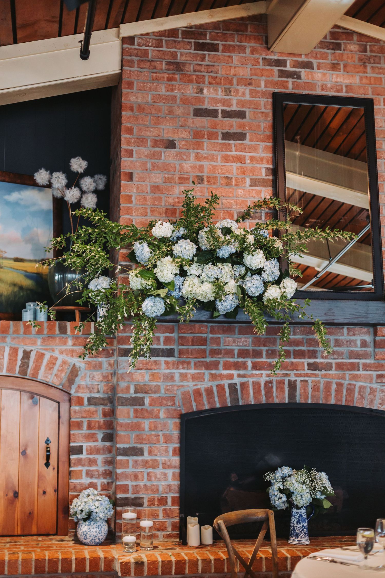 White and blue hydrangea floral arrangement displayed on rustic brick fireplace mantel at wedding venue.