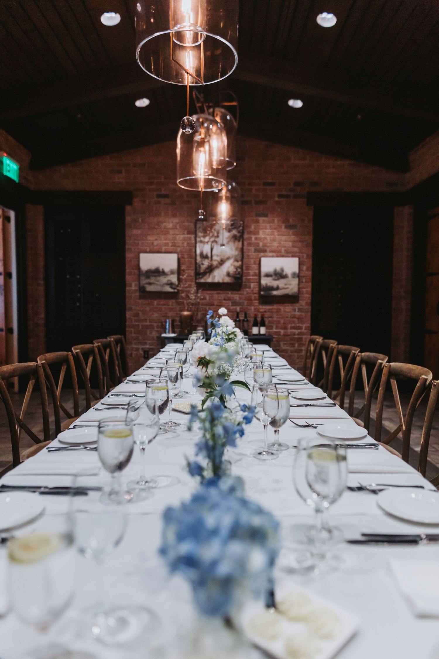 Elegant long wedding reception table with blue floral centerpieces and copper pendant lights in brick venue.