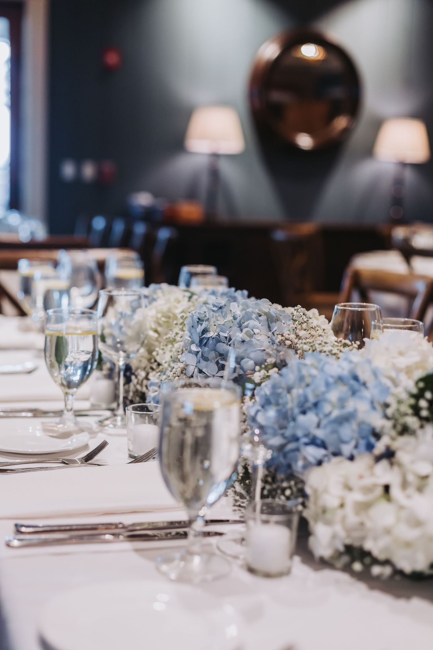 Wedding reception table set with blue and white hydrangea centerpieces, glassware, and white linens.