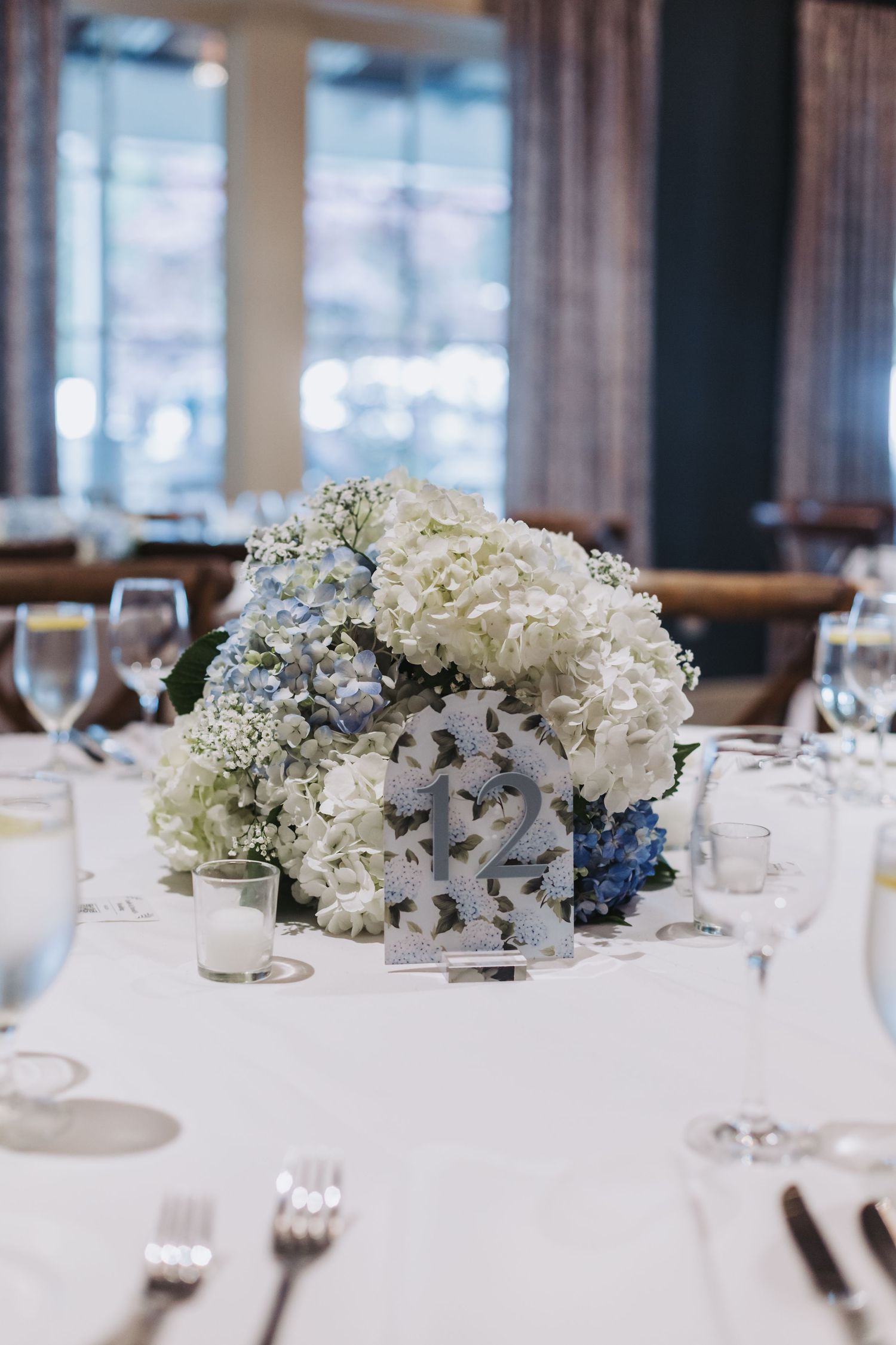 White and blue hydrangea centerpiece with table number 12 on white linen reception table.
