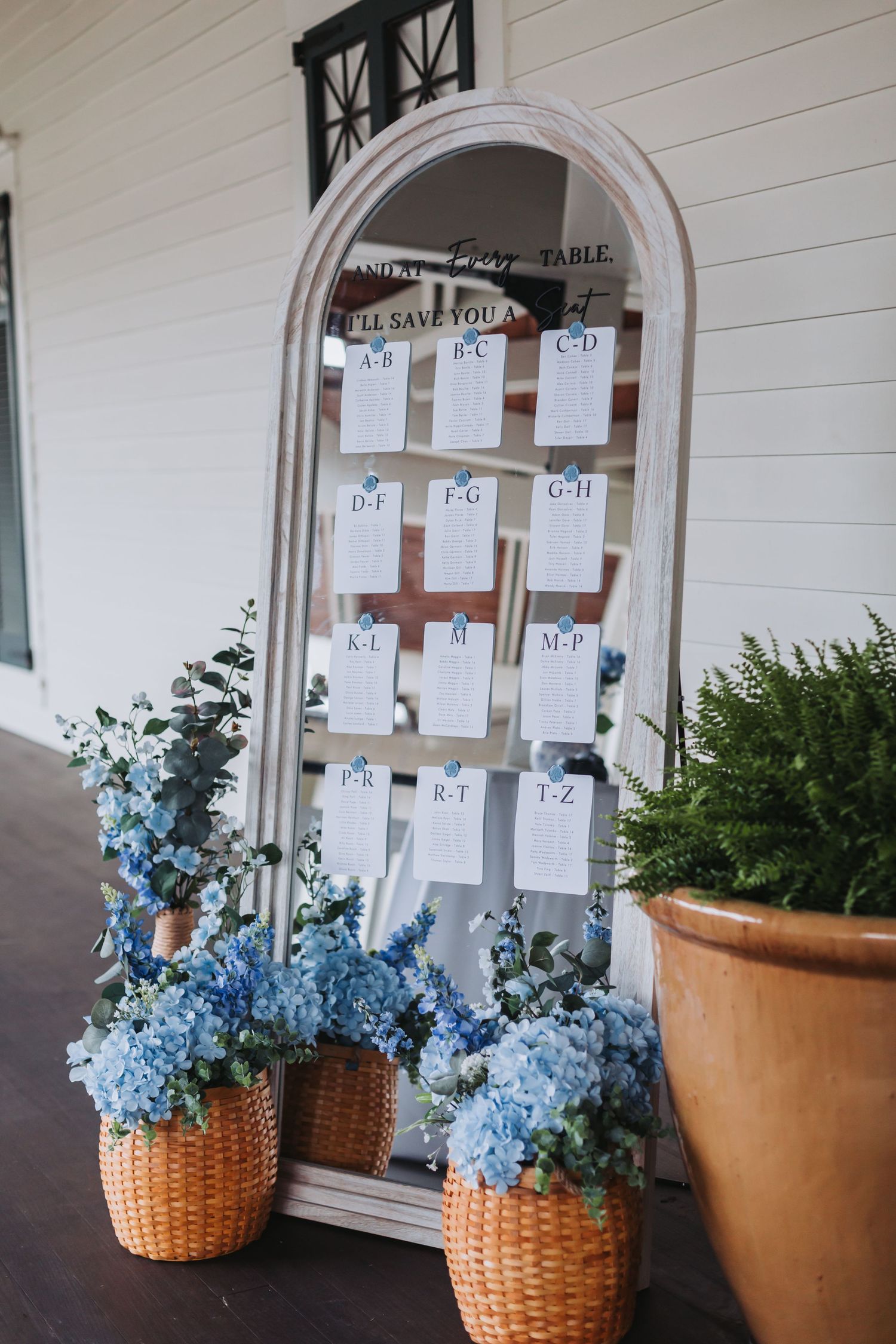 Wedding seating chart display on arched mirror decorated with blue hydrangeas in wicker baskets.